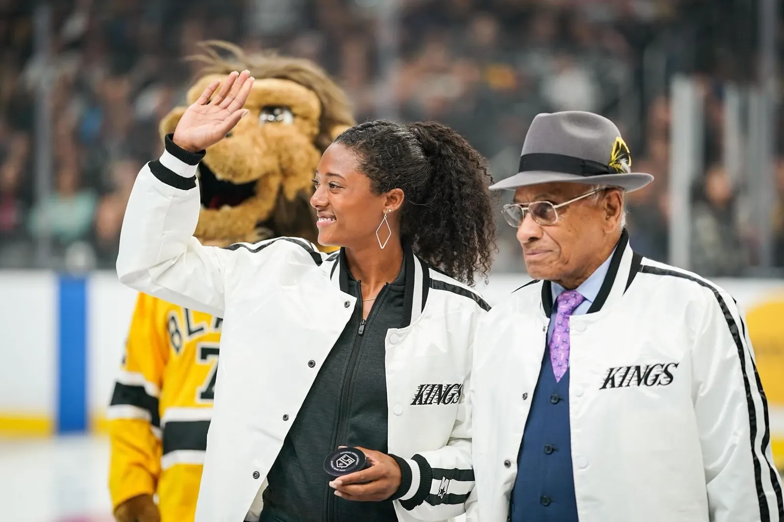 Blake Bolden in a white LA Kings jacket waving at a rink, beside Willie O'Ree and the Kings mascot