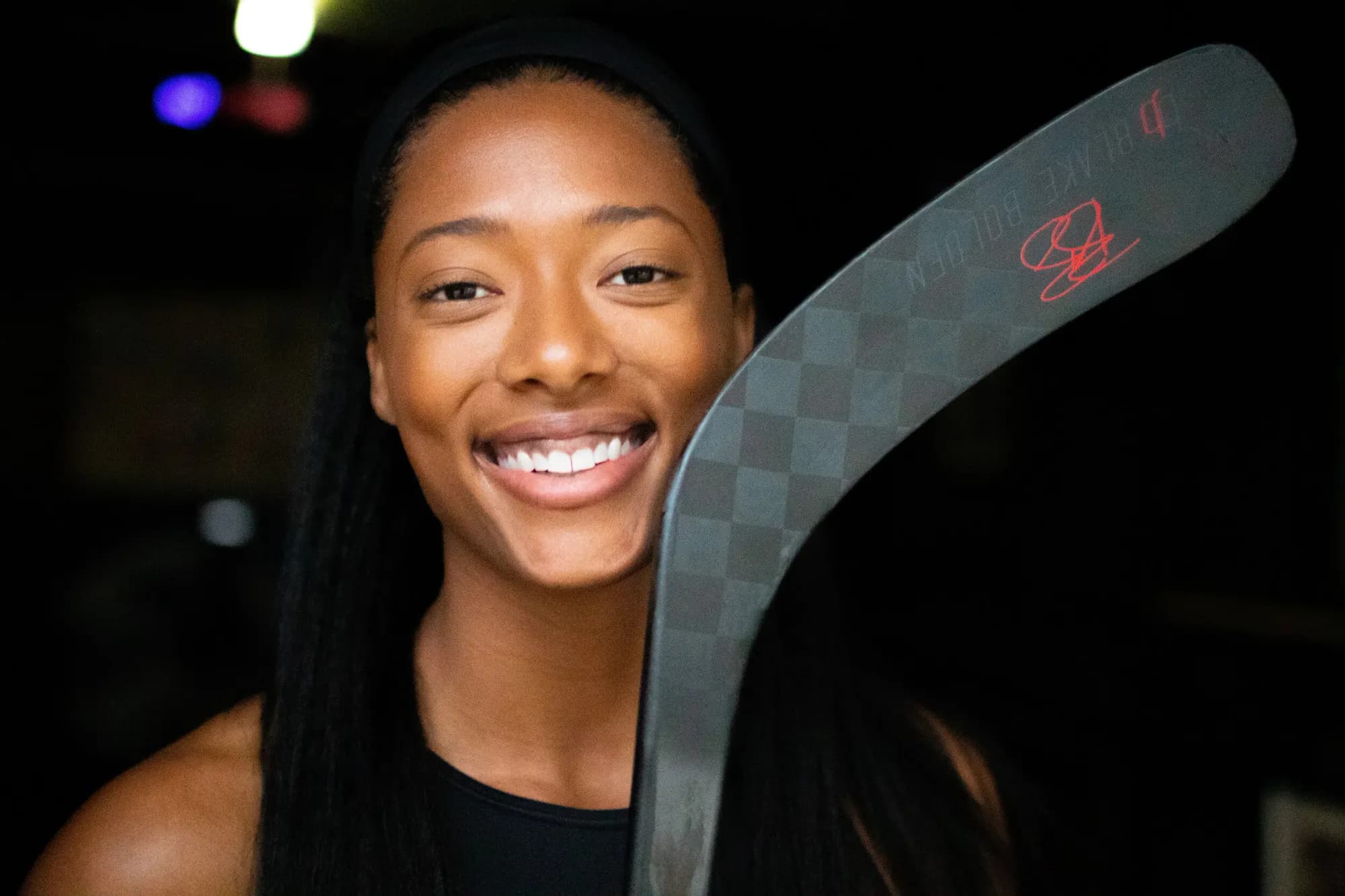 Blake Bolden smiling, a signed hockey stick resting on her shoulder